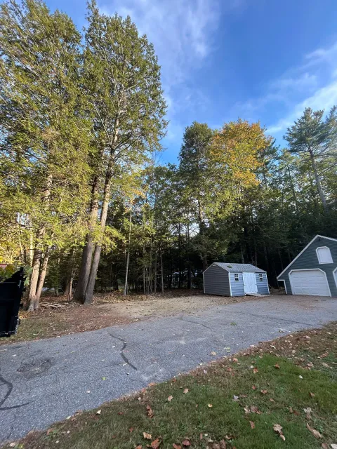 Gravel driveway beside tall trees, with stacked white materials and a small shed under a blue sky.