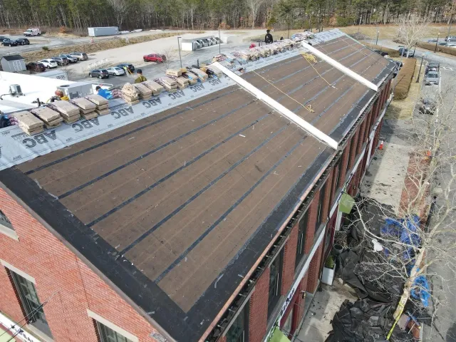Aerial view of a large brick building with a dark roof, surrounded by construction materials and equipment.