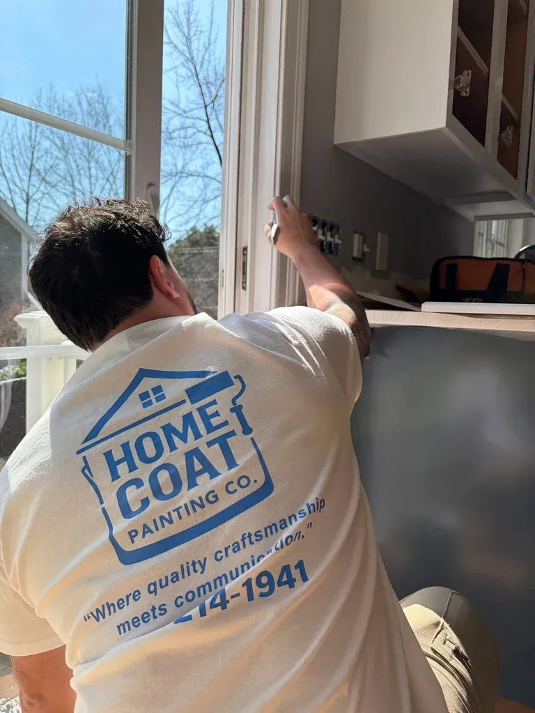Worker in Home Coat Painting Co. shirt standing at kitchen cabinets by a window