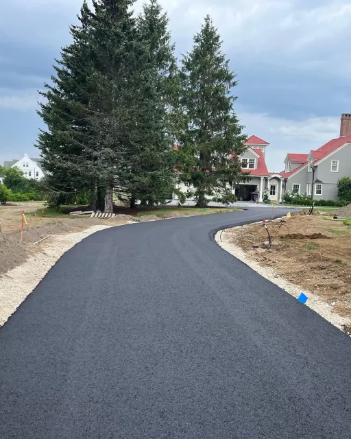 Newly paved asphalt driveway curves towards a house with tall trees in the background. Cloudy sky.