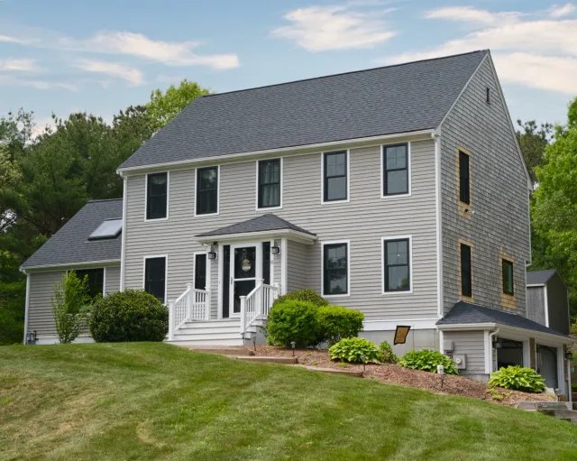 A large, multi-story house with beige siding, black window frames, and a dark gray roof, surrounded by a green lawn, shrubs, and trees with a partly cloudy sky in the background.