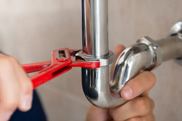 A person using red pliers to tighten a metal pipe connector on a chrome U-bend pipe.