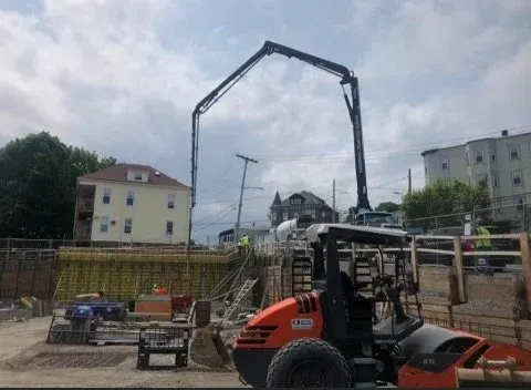 Construction site with a concrete pump truck pouring concrete into a foundation, with houses and trees in the background.