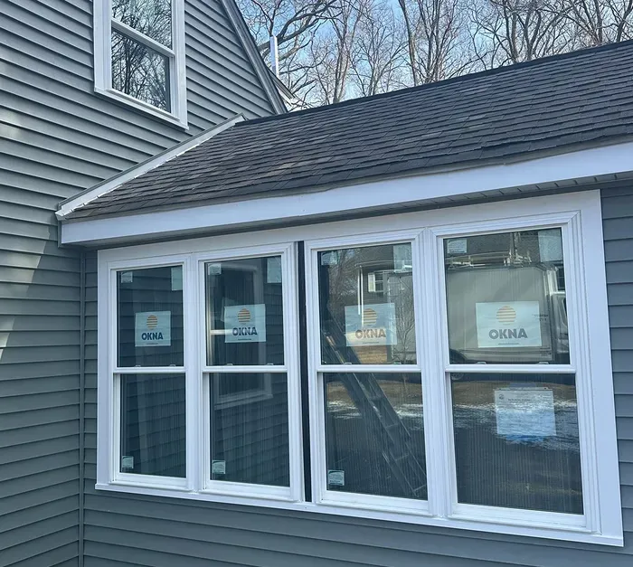 House exterior with white-framed windows and a dark shingled roof, partially covered in protective wrapping.