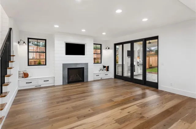 Bright living room with hardwood floors, fireplace, wall TV, stairs, and black French doors to a patio