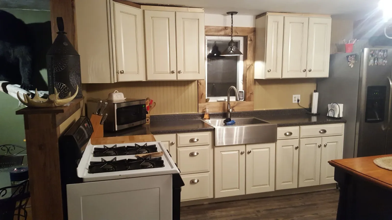 Kitchen with white cabinets, black countertops, stainless steel sink, microwave, gas stove, refrigerator, and various small appliances. There is a window above the sink and a hanging lamp. Part of a wooden dining table is visible in the foreground.