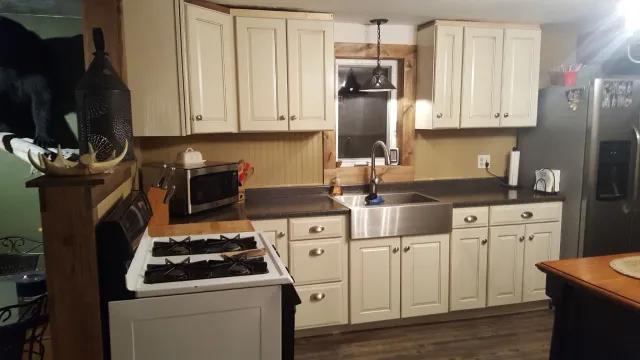 Kitchen with white cabinets, black countertops, stainless steel sink, microwave, gas stove, refrigerator, and various small appliances. There is a window above the sink and a hanging lamp. Part of a wooden dining table is visible in the foreground.