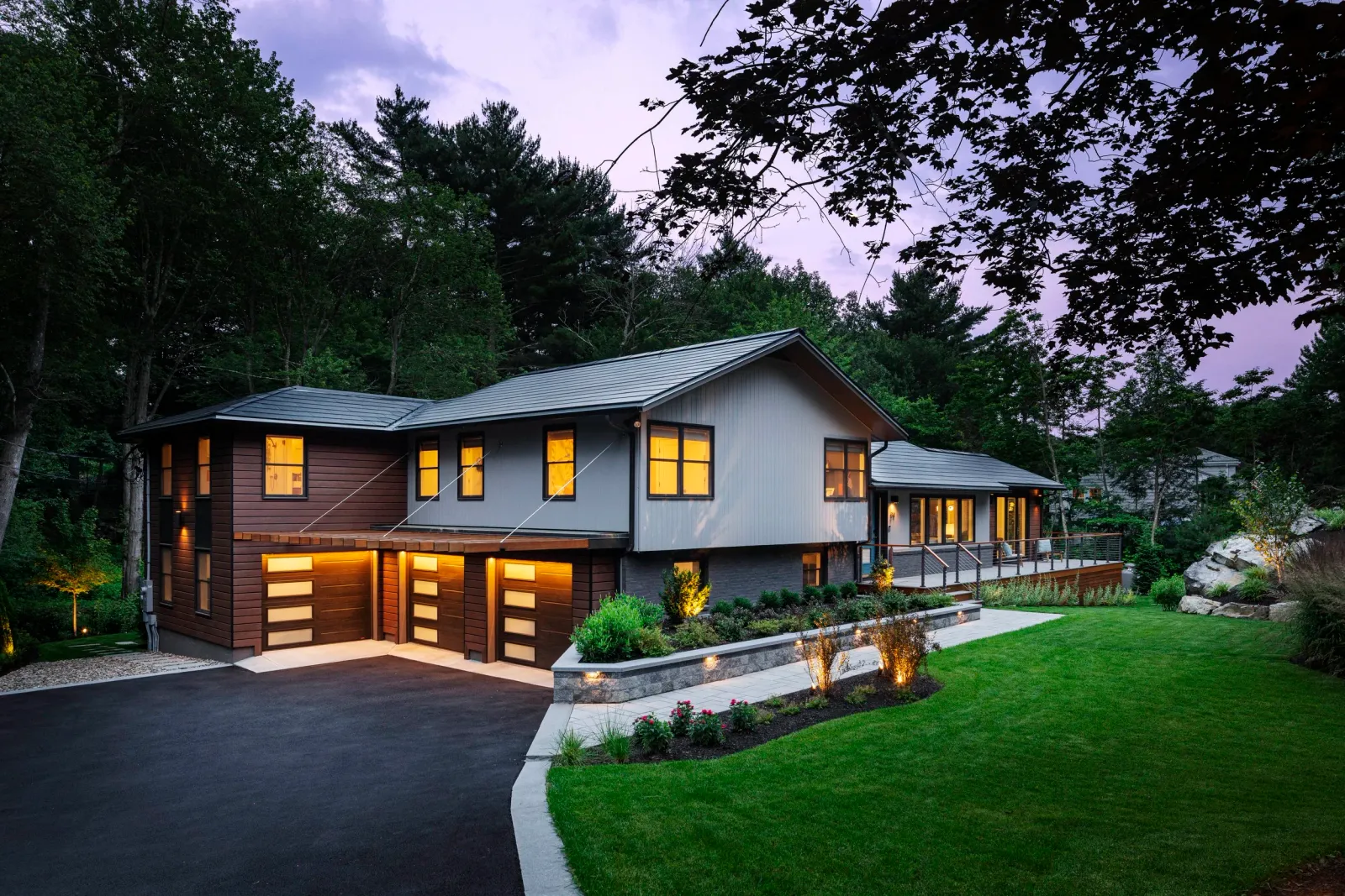 Modern two-story house with a sloped gray roof, a mix of gray and brown exterior walls, a driveway, a landscaped front yard with green grass, bushes, and flowers, and surrounded by trees.