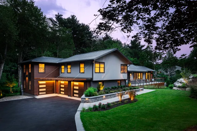 Modern two-story house with a sloped gray roof, a mix of gray and brown exterior walls, a driveway, a landscaped front yard with green grass, bushes, and flowers, and surrounded by trees.