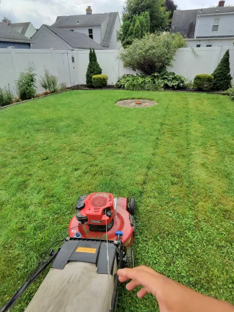 View from a lawnmower operator's perspective, looking towards a green lawn with garden beds and shrubs, enclosed by a white fence, with residential houses in the background under cloudy sky.