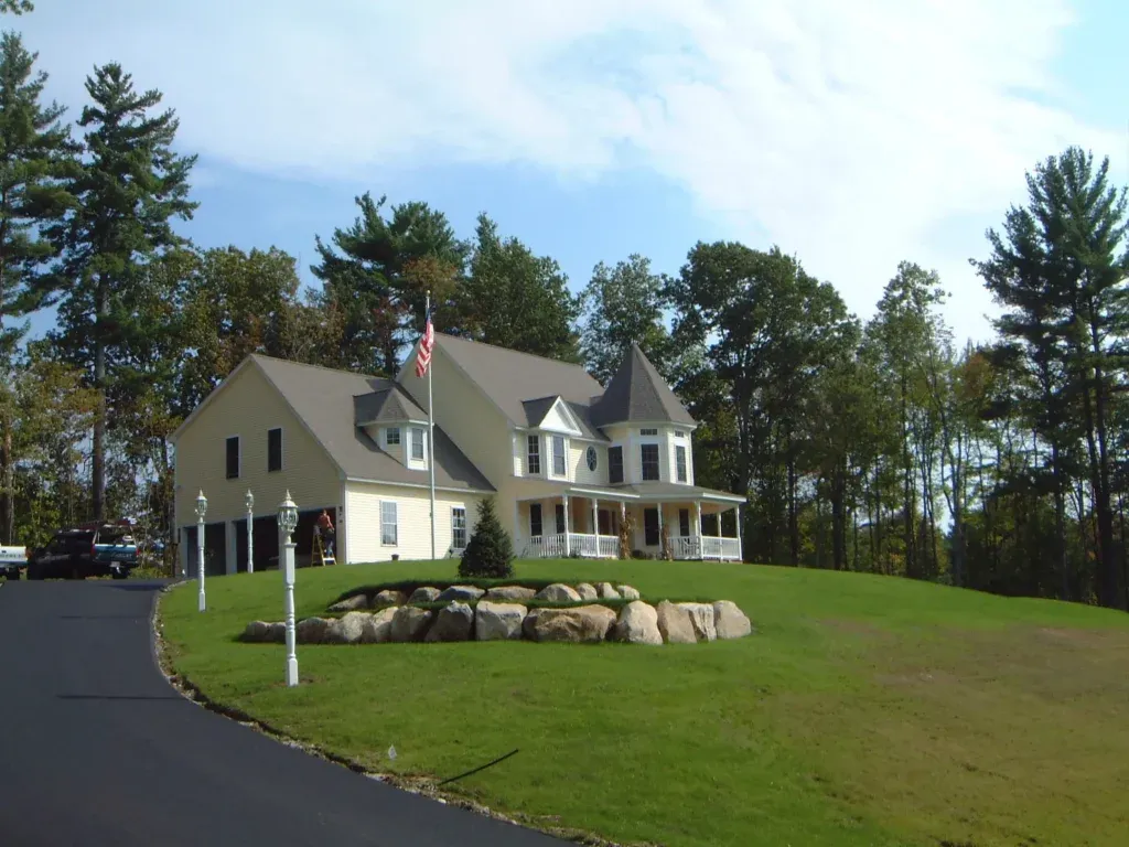 Large two-story colonial home with wrap-around porch, flagpole, and boulder landscaping on a green lawn