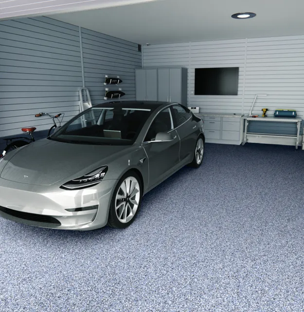 A modern garage with a silver electric car parked inside. The garage has light gray paneled walls, a blue carpeted floor, and a ceiling light. There are some tools and storage cabinets against the wall, along with a flat-screen TV mounted on the wall.
