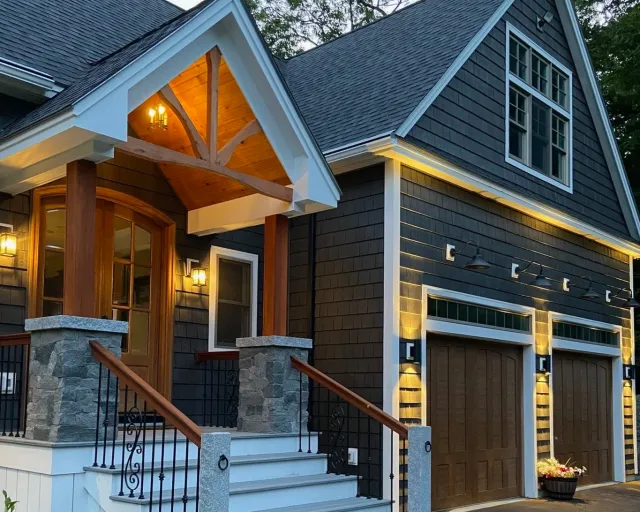Exterior of a modern house with a dark shingle facade, stone pillars at the front entrance, wooden handrails, a double garage door, and outdoor lighting, during twilight.