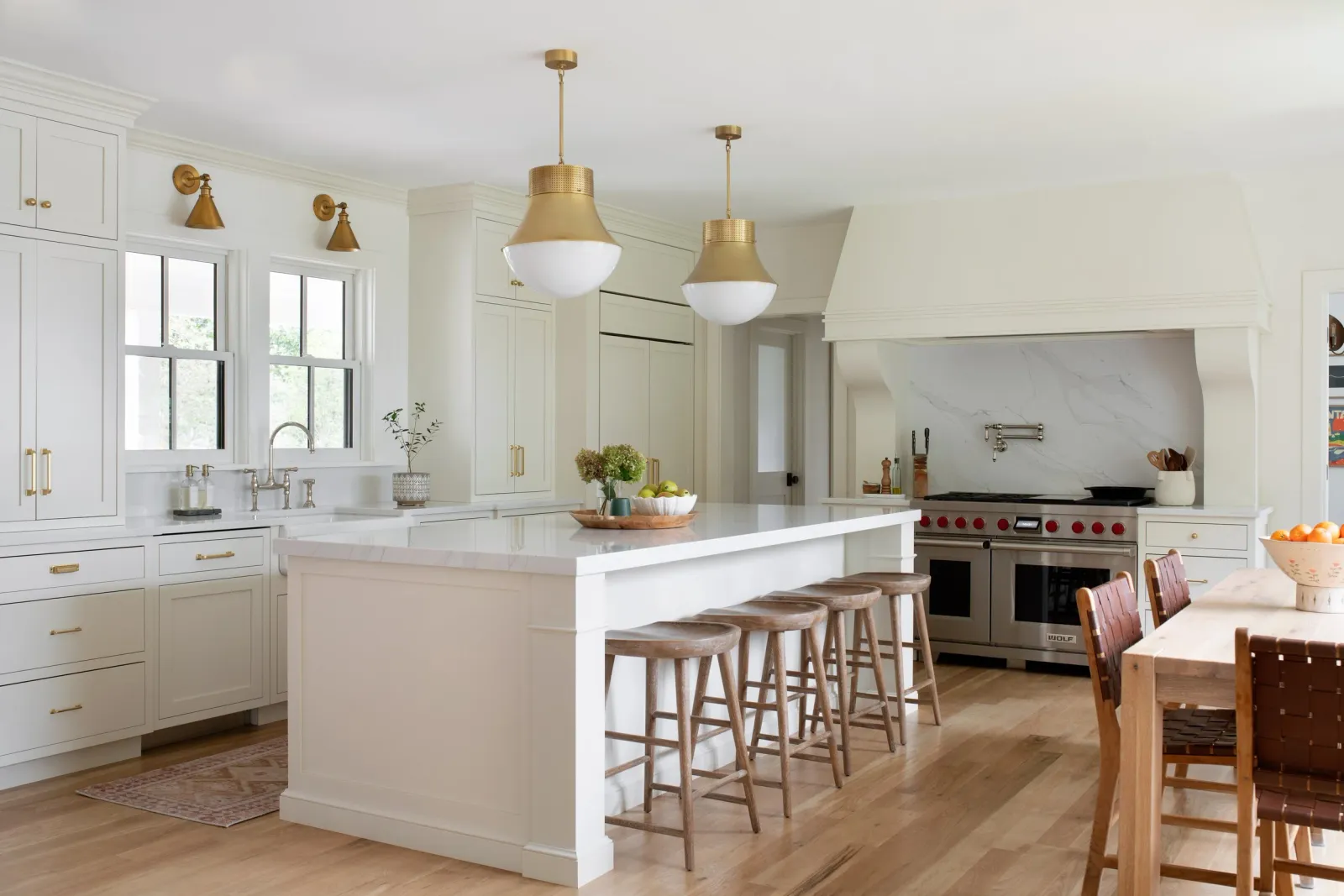 Modern white kitchen with a large island, wooden stools, gold pendant lights, stainless steel appliances, and a window above the sink.
