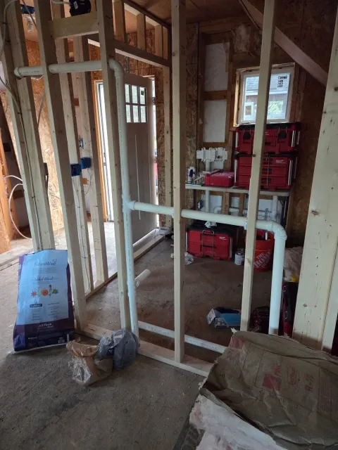 Interior of a house under construction with exposed wooden framing, PVC piping, a window, tools, and construction materials.