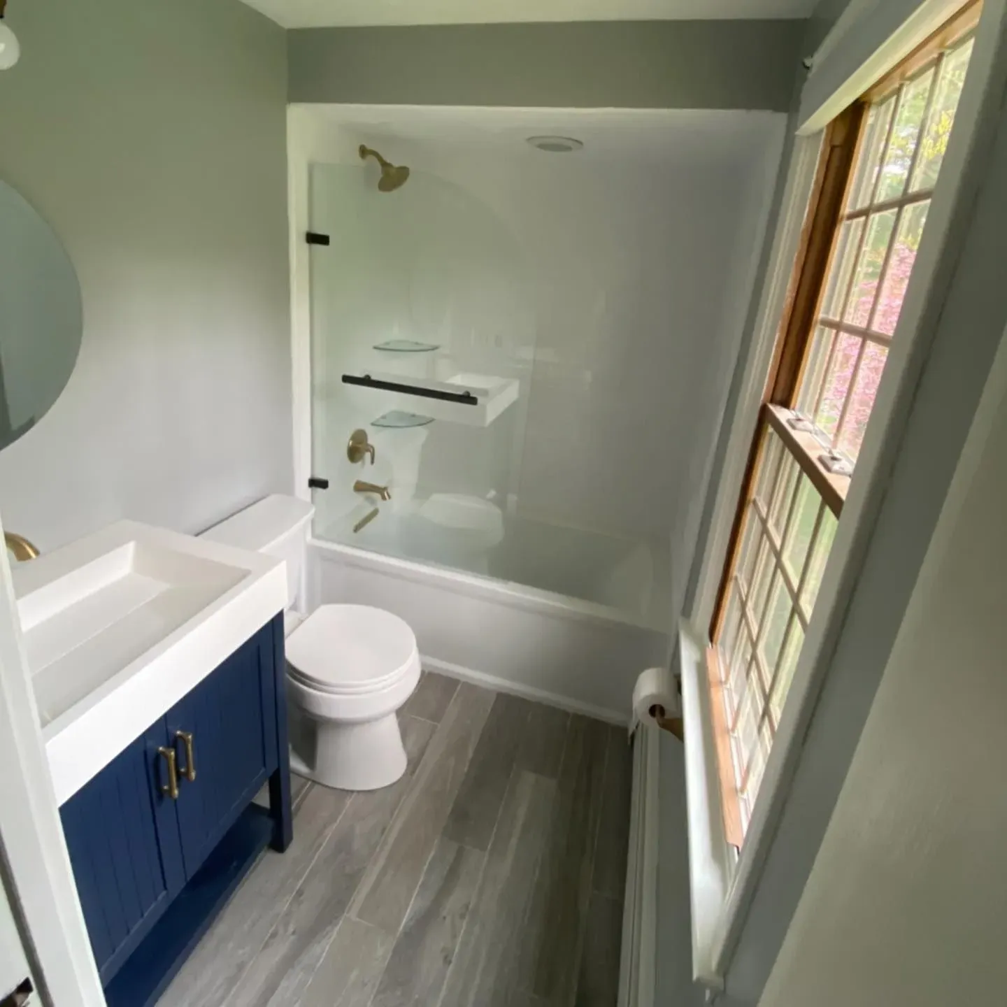 A small, modern bathroom featuring a blue vanity, a toilet, and a white bathtub-shower combo with glass shelving.