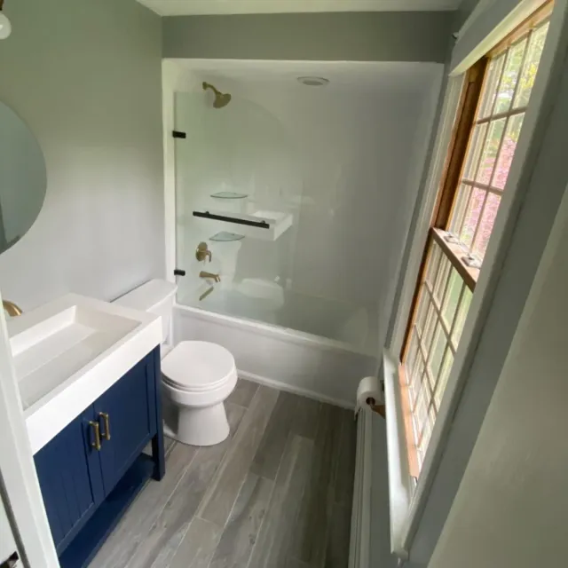 A small, modern bathroom featuring a blue vanity, a toilet, and a white bathtub-shower combo with glass shelving.