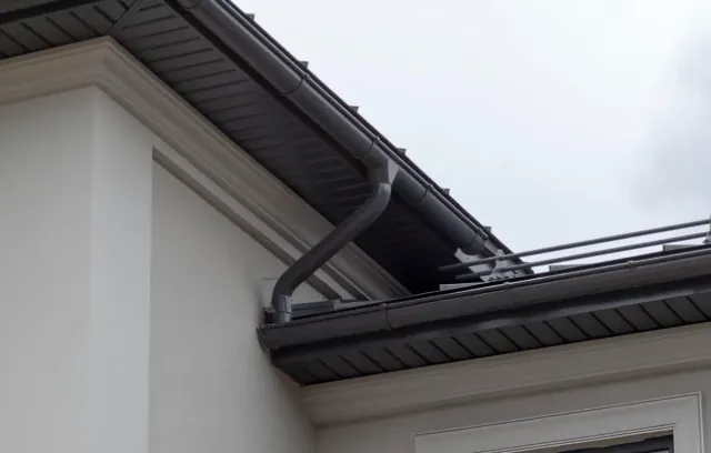 Close-up of a house corner showing a dark-colored gutter and downspout system attached under the roof eaves.