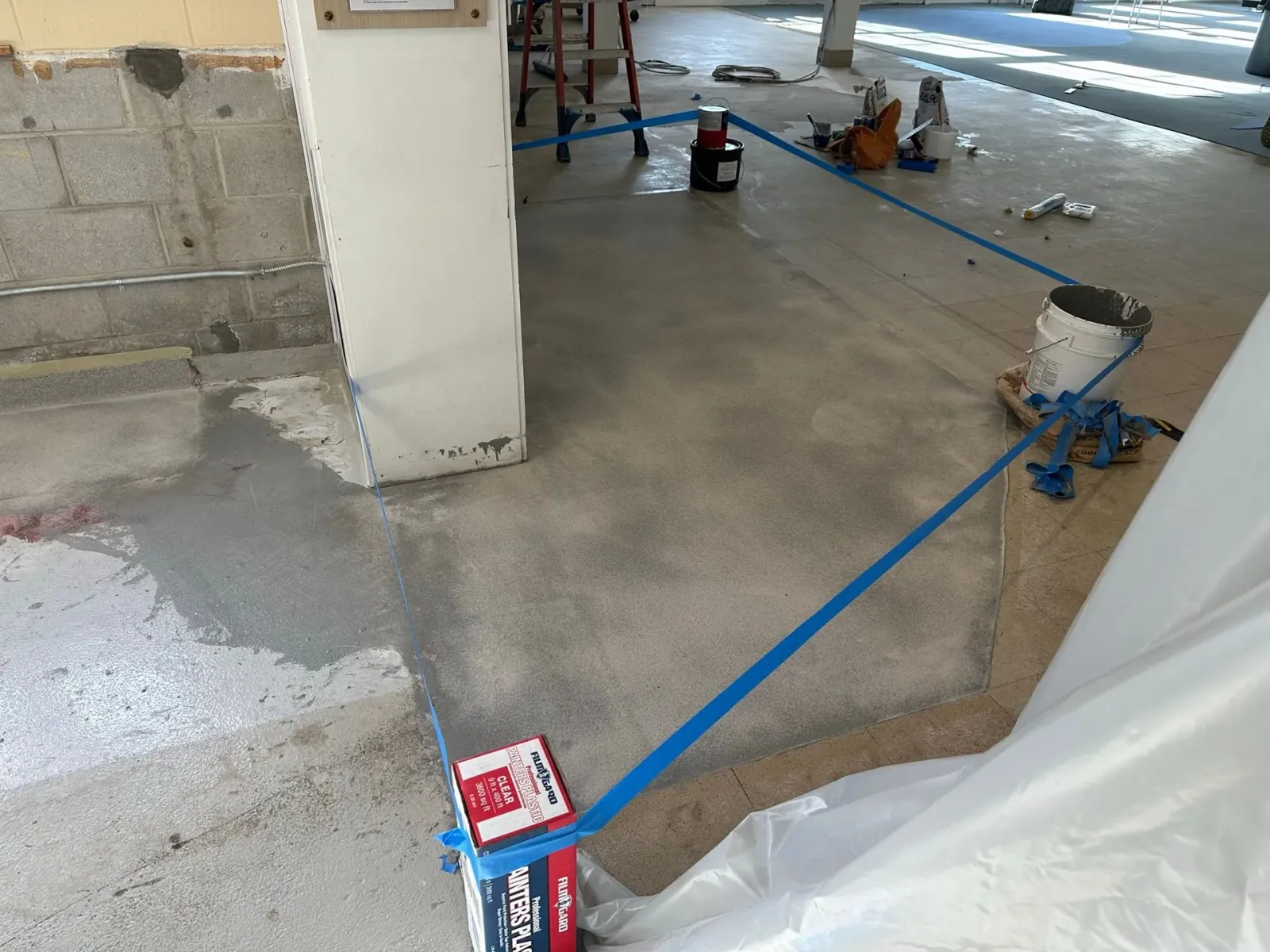 Blue tape lines a construction floor with pillars, tools, and a plastic sheet in an unfinished room