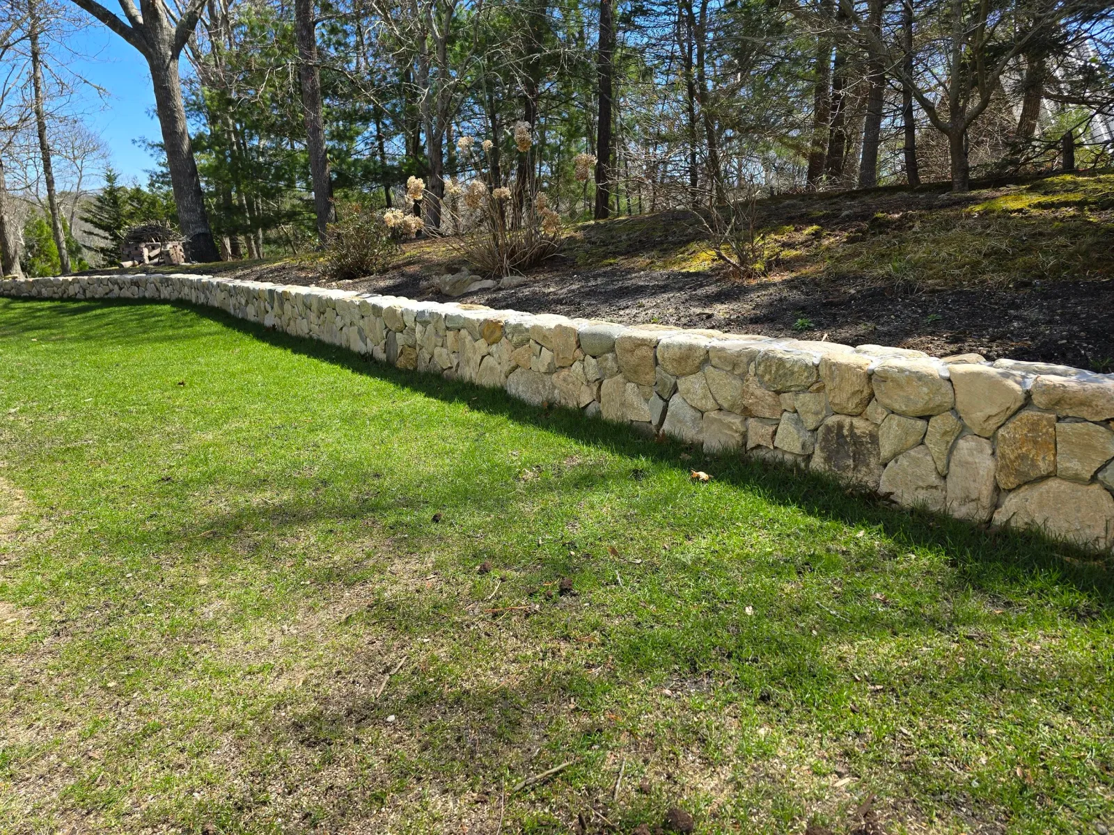 A stone retaining wall separates a well-maintained grassy lawn from a wooded area with trees and bushes.
