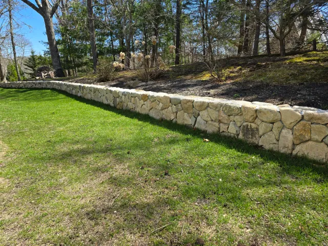 A stone retaining wall separates a well-maintained grassy lawn from a wooded area with trees and bushes.