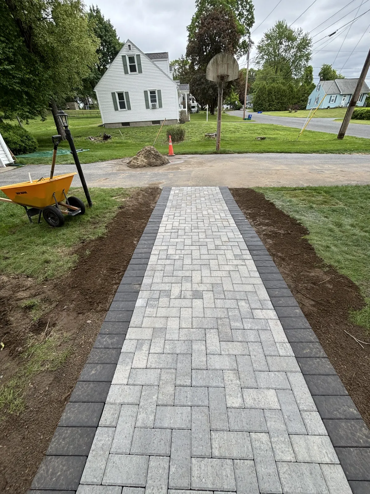 A newly paved pathway with light gray and dark gray bricks, bordered by dark gray bricks, leading to a residential street with houses, trees, and a basketball hoop in the background, surrounded by grass and construction materials.