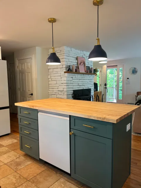 Kitchen island with a light wood countertop, green cabinets with gold handles, and a white appliance in the middle. Black pendant lights hanging from the ceiling above.