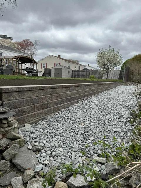 A landscaped backyard featuring a stone retaining wall bordered by a bed of light gray gravel under a cloudy sky.
