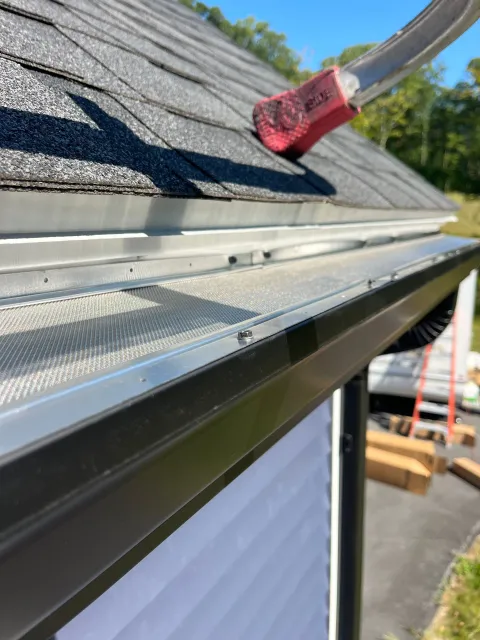Red brush cleaning debris from a roof gutter beside gray shingles.