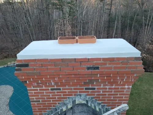 Brick chimney with a white cap and two bricks on top, set against trees outdoors