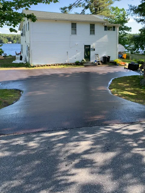 A newly paved driveway in front of a white house with a lake in the background, surrounded by trees.