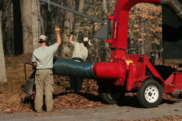 Two workers loading leaves into a red wood chipper beside a tree-lined road.