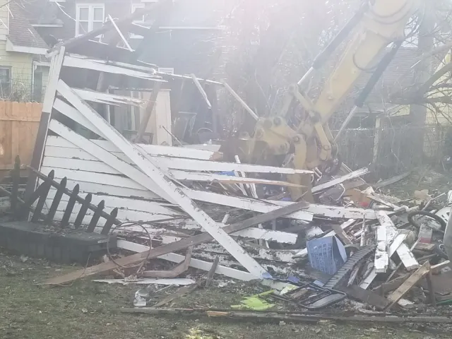 Debris from a collapsed or destroyed house, including a disassembled fence, scattered wood, a blue plastic crate, and a yellow excavator arm in the background.