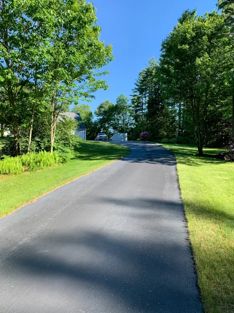 A curved asphalt driveway in a residential area surrounded by green grass, trees, and shrubs, with a house and parked car visible in the background under a clear blue sky.