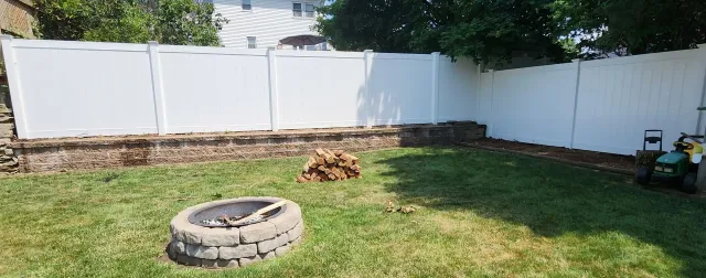 Backyard with white fence, grassy lawn, fire pit ring, and a small brush pile near the back fence.