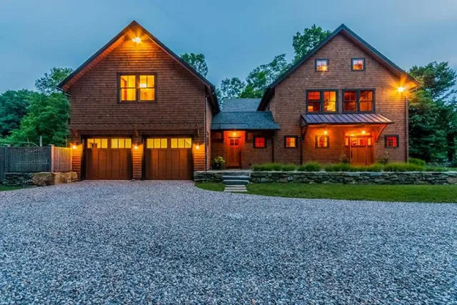 A large two-story wooden house with a gravel driveway, illuminated warm lights, multiple windows, a front porch, and surrounded by trees at dusk.