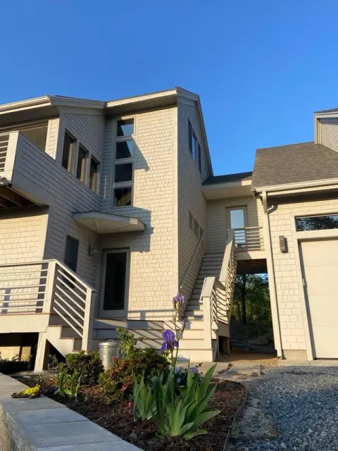 Modern beige townhouse exterior with stairs, balconies, and a flower bed under a blue sky