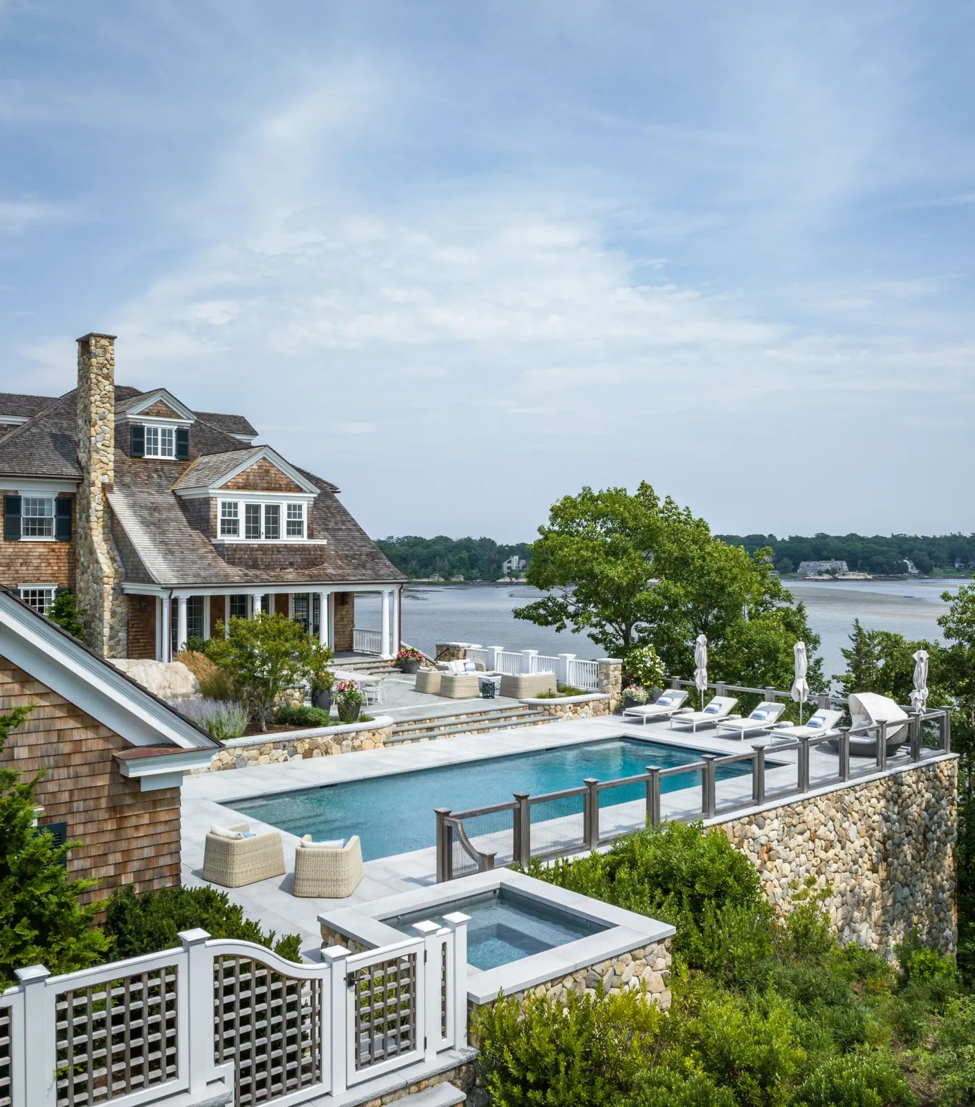 A view of a waterfront house with a crystal-clear swimming pool, sparkling hot tub, and aesthetic relaxing lounge area built on a larger stone base surrounded by lush verdant greenery, overlooking a river under a partly cloudy sky.