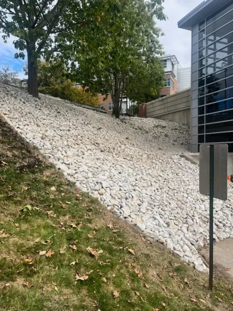 White gravel embankment beside a building, with trees and a sloped grassy edge in front.