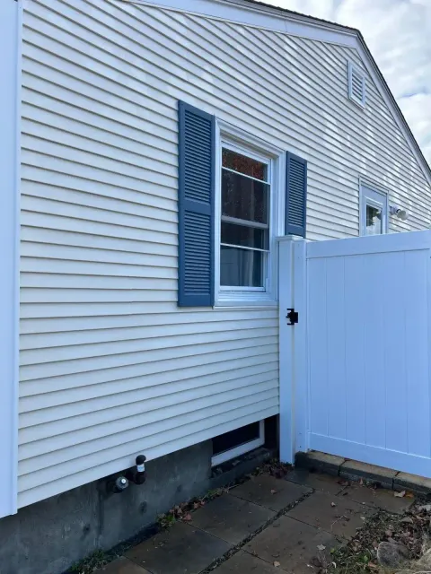 Exterior side wall of a house with white vinyl siding, a window with blue shutters, and a white gate on a paved pathway.