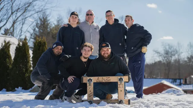 Group of seven men pose outdoors in the snow, some kneeling, holding a wooden structure; sunny day.
