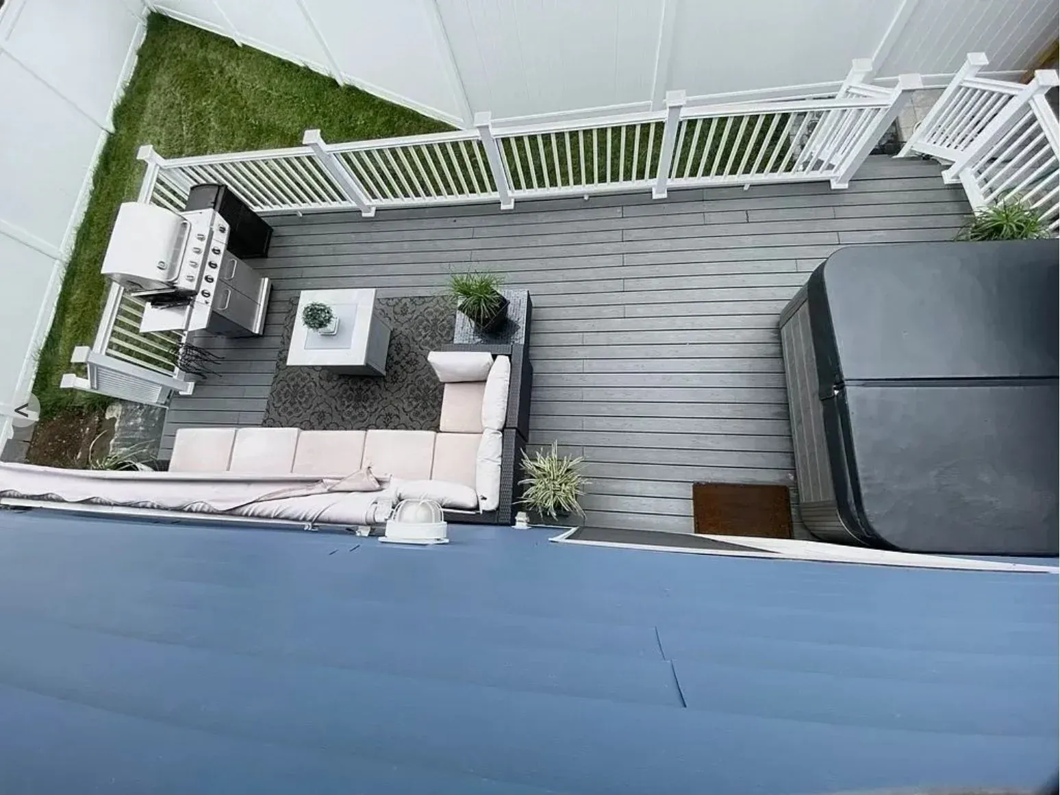 Aerial view of a backyard patio with a gray deck, white railing, L-shaped outdoor sofa, coffee table, a gas grill, hot tub, and greenery.