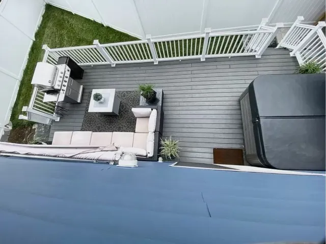 Aerial view of a backyard patio with a gray deck, white railing, L-shaped outdoor sofa, coffee table, a gas grill, hot tub, and greenery.