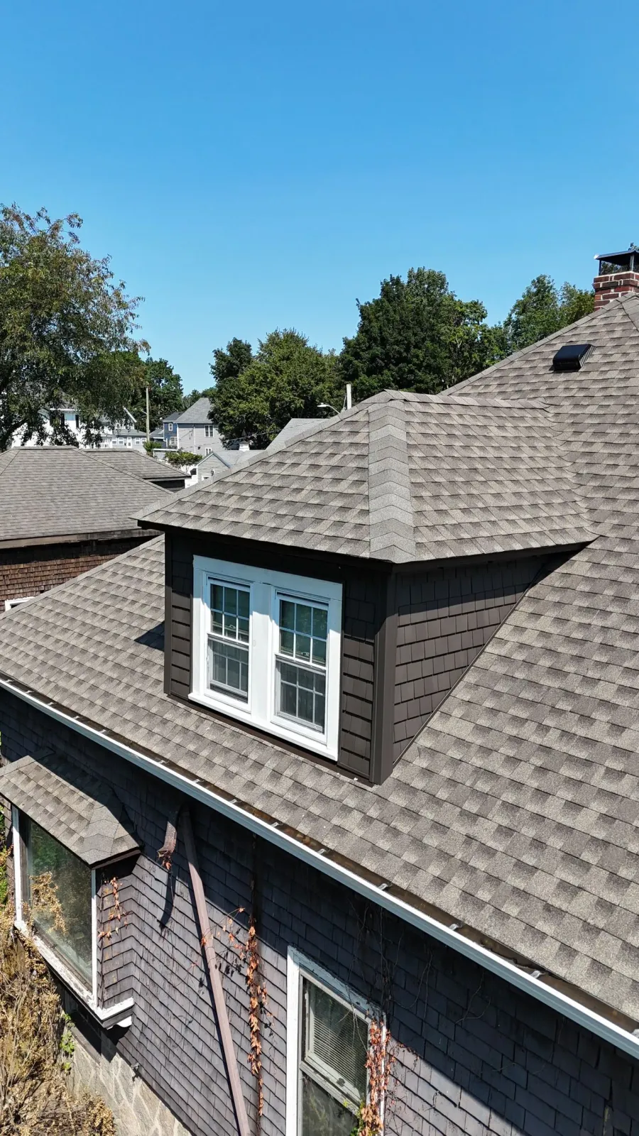 A dormer window with white trim on a house with dark gray shingles and a gabled roof under a clear blue sky.