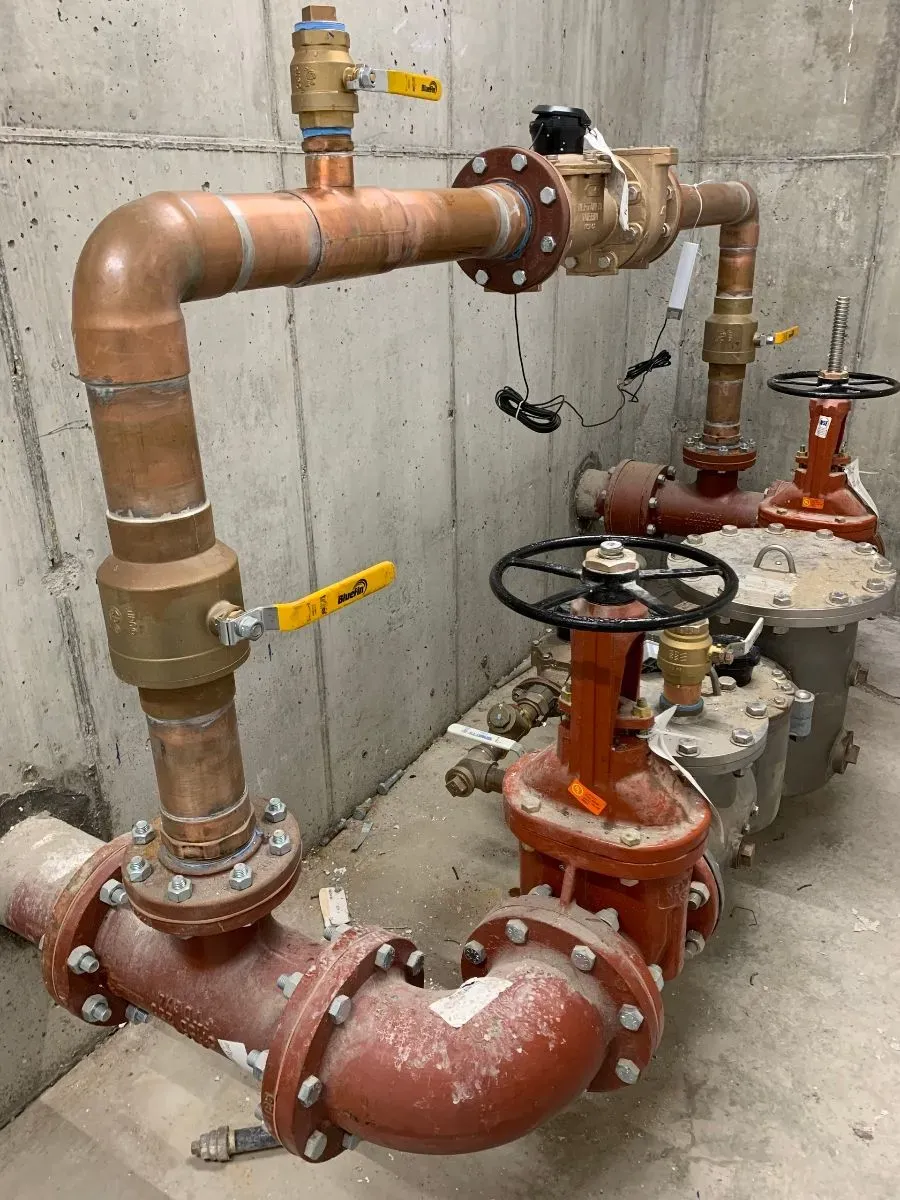 A complex plumbing system with copper and iron pipes, valves with black handwheels, yellow handles, and various fittings installed against a concrete wall in an industrial setting in Middlesex and Suffolk County, MA.