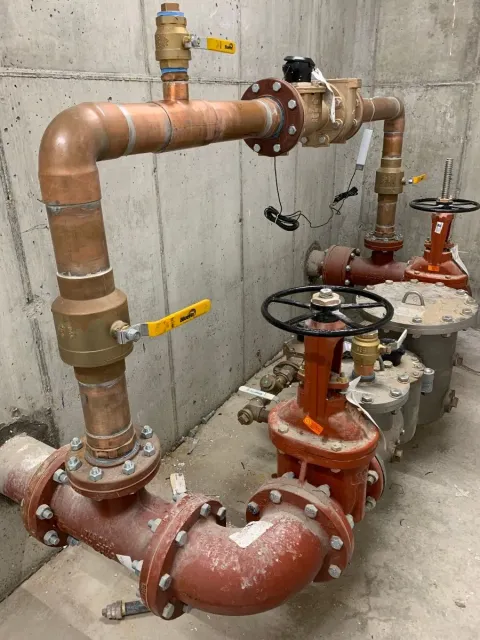 A complex plumbing system with copper and iron pipes, valves with black handwheels, yellow handles, and various fittings installed against a concrete wall in an industrial setting in Middlesex and Suffolk County, MA.