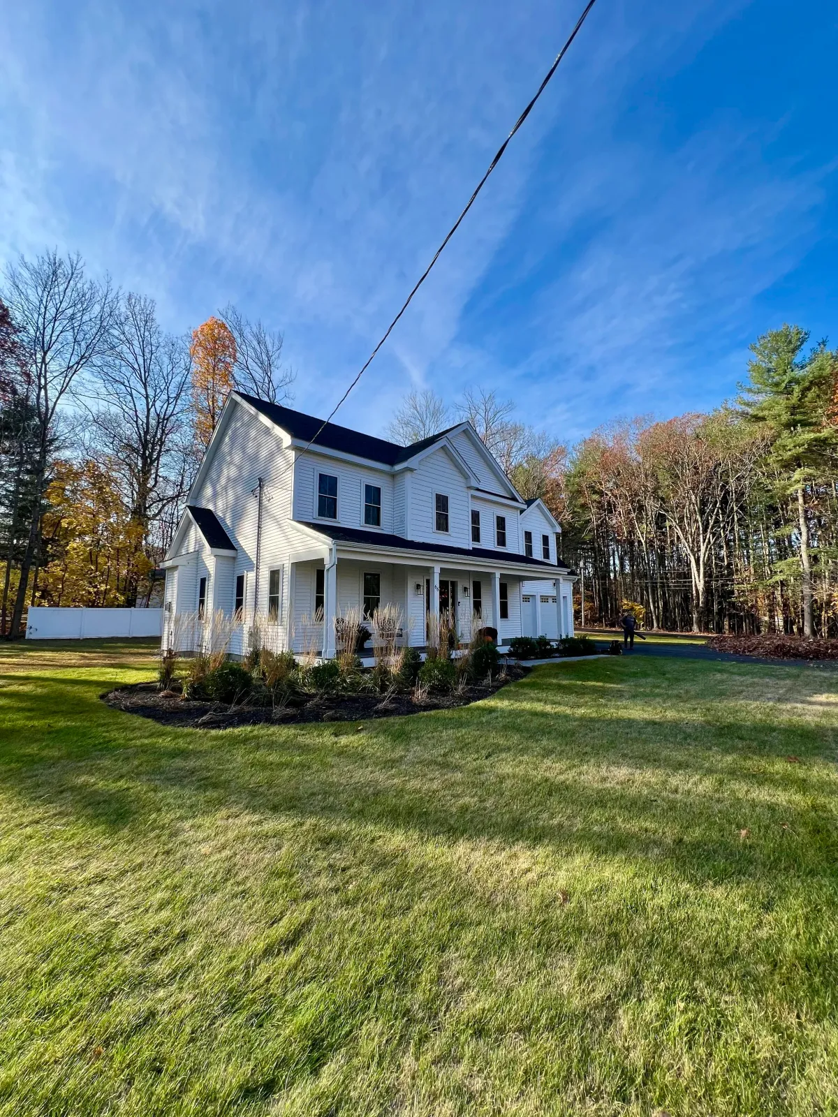White two-story house with a front porch, black roof, and multiple windows, surrounded by a well-maintained lawn and trees with autumn leaves, under a clear blue sky.