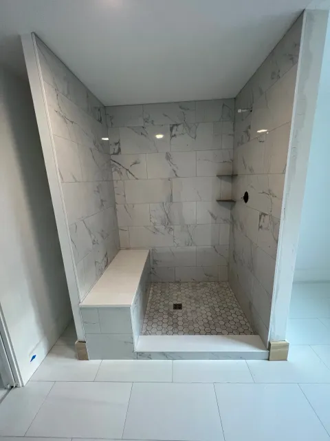 A modern walk-in shower with marble tile walls, built-in bench, and hexagonal floor tiles, with two small stainless steel shelves and a drain on the shower floor.