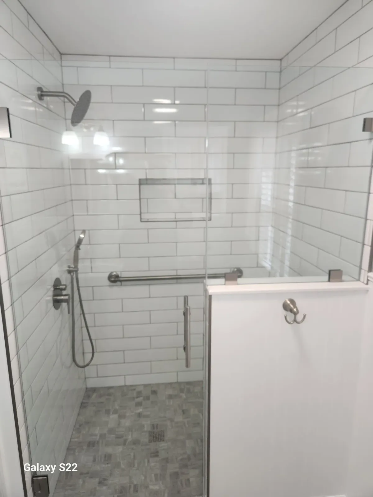A walk-in shower with white subway tile walls, a rain showerhead, a handheld shower, a narrow glass door with a metal handle, a wall-mounted grab bar, a small wall-mounted shelf, and a dark gray tiled floor with a drain.