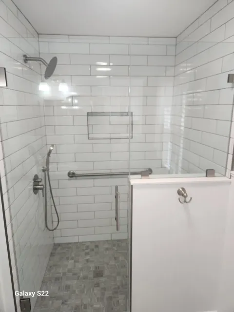 A walk-in shower with white subway tile walls, a rain showerhead, a handheld shower, a narrow glass door with a metal handle, a wall-mounted grab bar, a small wall-mounted shelf, and a dark gray tiled floor with a drain.